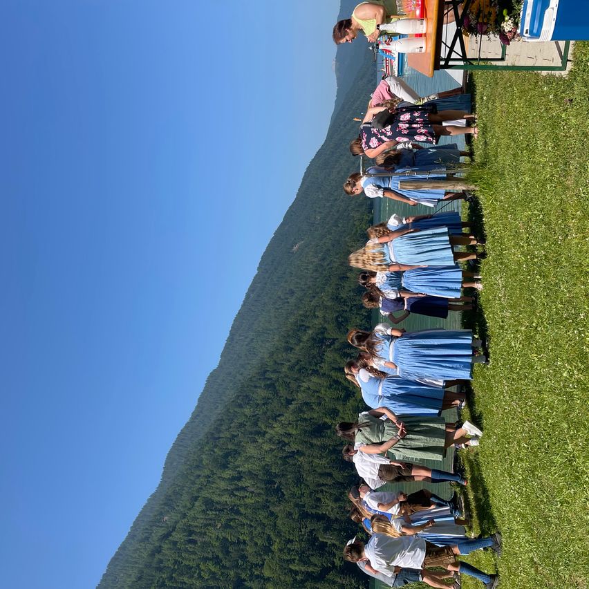 Eine Gruppe von Menschen in traditioneller Kleidung steht auf einem grasbewachsenen Hügel, mit einem See und Bergen im Hintergrund. Es gibt auch einige Leute, die in der Nähe eines Tisches mit Flaschen und Blumen stehen.