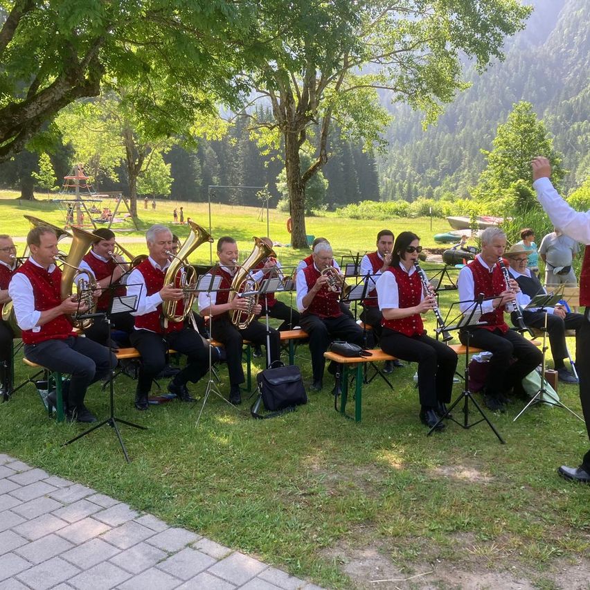 Ein Blasorchester spielt auf einer Rasenfläche vor einem Bergpanorama. Die Musiker sitzen auf Bänken und spielen ihre Instrumente.
