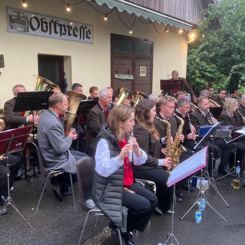 Eine Blechbläsergruppe spielt im Freien vor einem Gebäude mit dem Schild 'Oberprellle'. Die Musiker sitzen und spielen verschiedene Instrumente, darunter Trompeten und Saxophone. Vor ihnen stehen Flaschen und ein Notenständer.