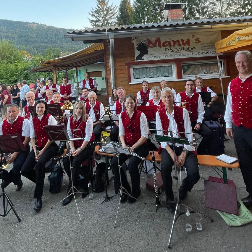 Eine Gruppe von Musikern in roten Westen posiert für ein Foto vor Manu's Friseurstuble, mit einer Bergkette im Hintergrund.