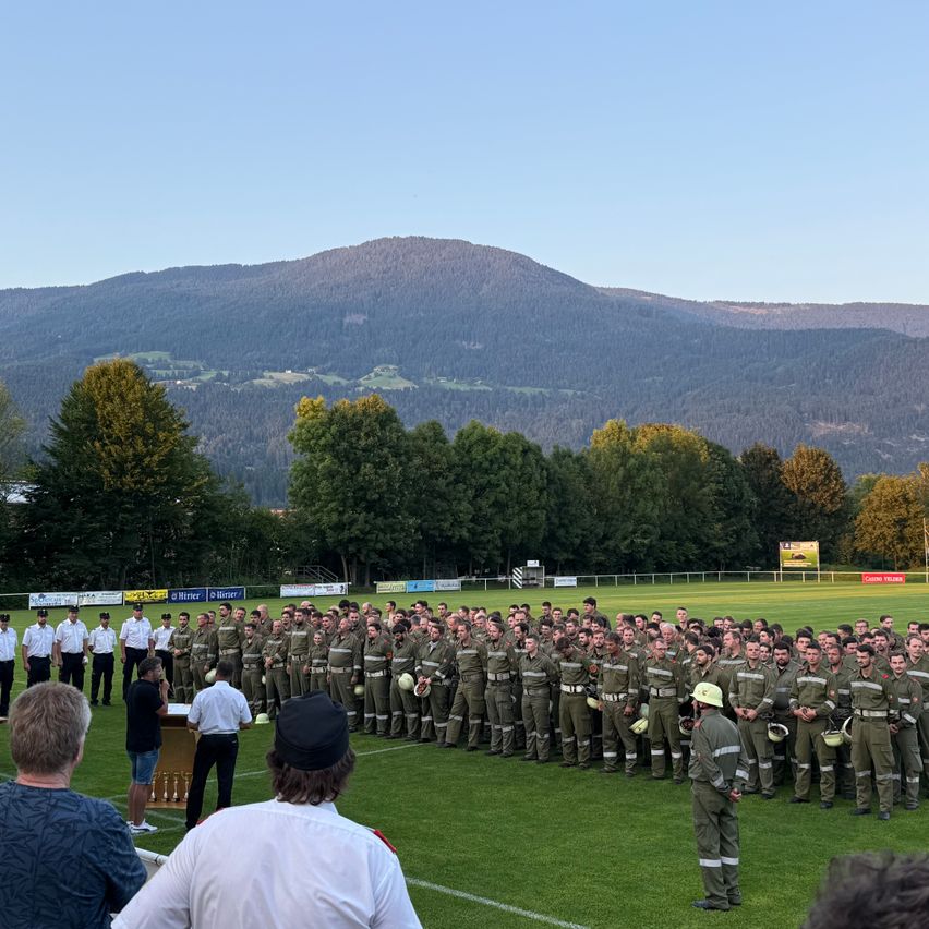Eine Gruppe von Personen in Uniform steht auf einem Feld mit Bergen im Hintergrund. Zuschauer beobachten die Veranstaltung.