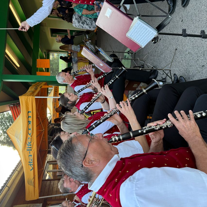 Eine Gruppe von Musikern in roten Westen spielt ihre Instrumente. Der Mann vorne spielt Klarinette, während die anderen Trompete und Posaune spielen. Sie sind wahrscheinlich bei einer Veranstaltung im Freien aufgetreten.
