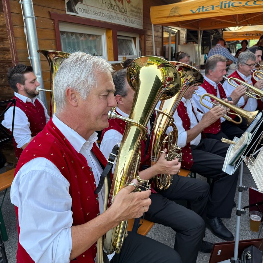 Eine Gruppe von Musikern in roten Westen spielt Blechblasinstrumente bei einer Veranstaltung im Freien. Ein Mann vorne spielt Tuba, während andere Trompeten spielen. Sie sitzen auf einer Bank unter einem Zelt.