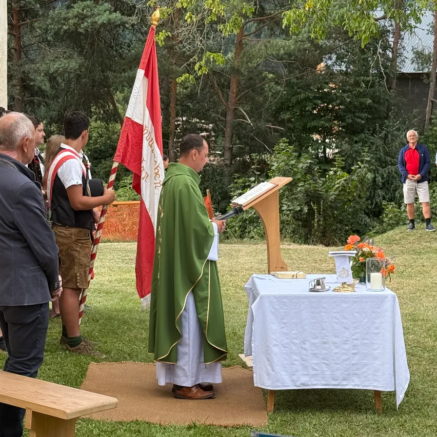 Ein Priester steht an einem Podium und liest aus einem Buch vor, mit einer Flagge und Menschen, die sich hinter ihm auf einer Grasfläche versammelt haben.