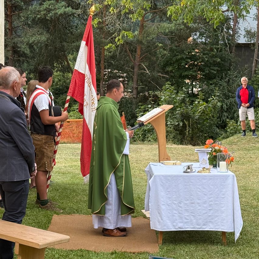 Ein Priester steht an einem Podium und liest aus einem Buch vor, mit einer Flagge und Menschen, die sich hinter ihm auf einer Grasfläche versammelt haben.