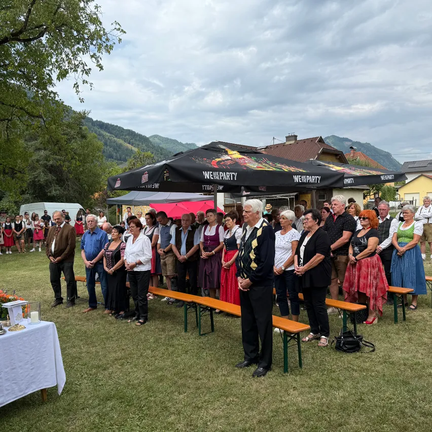 Auf einem Außenbereich steht eine Gruppe von Menschen vor einem großen Zelt. Das Zelt hat ein Baldachin mit den Worten 'Wir feiern' darauf geschrieben. Ein Tisch mit einem weißen Tuch und einer Kerze steht auf der linken Seite. Im Hintergrund sind Berge und Häuser zu sehen.