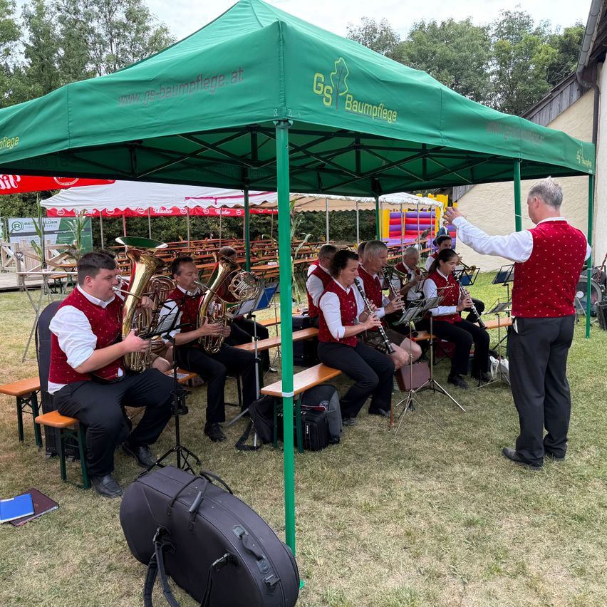 Eine Gruppe von Musikern spielt verschiedene Instrumente unter einem grünen Zelt. Das Zelt hat eine Website darauf gedruckt. Die Musiker sitzen auf Bänken, während sie spielen.