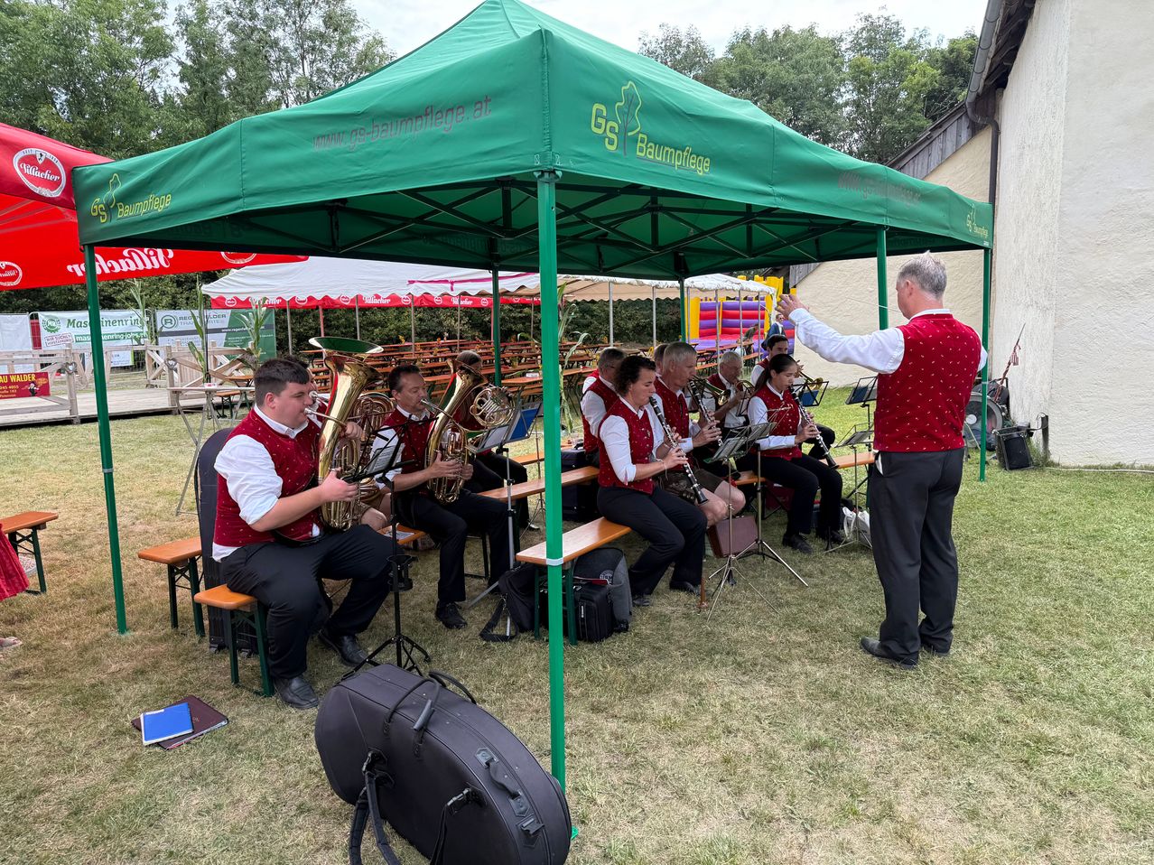 Eine Gruppe von Musikern spielt verschiedene Instrumente unter einem grünen Zelt. Das Zelt hat eine Website darauf gedruckt. Die Musiker sitzen auf Bänken, während sie spielen.