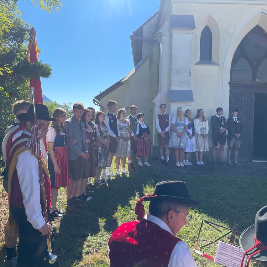 Eine Gruppe von Menschen in traditioneller Kleidung steht vor einer Kirche. Einige tragen Hüte, Schuhe und eine Fahne. Eine Person im Vordergrund trägt eine Brille und eine Weste.