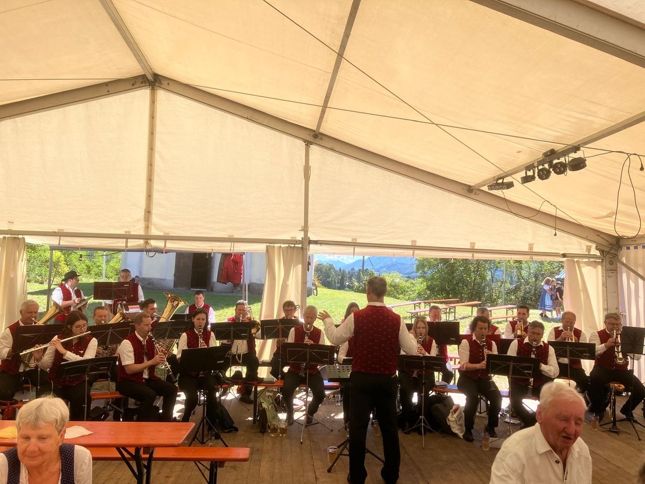Eine Band spielt unter einem weißen Zelt, mit einem Mann vorne, der sie leitet. Sie spielen verschiedene Instrumente, darunter Trompeten, Posaunen und Klarinetten. Das Publikum sitzt auf Stühlen, und einige stehen. Im Hintergrund gibt es Bäume und ein Gebäude.