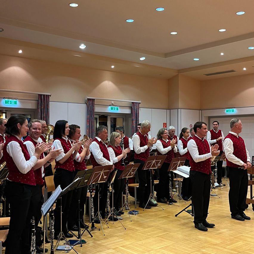 Eine Gruppe von Musikern in roten Westen und schwarzen Hosen spielt drinnen. Sie halten Instrumente und stehen vor Notenständern. Einige klatschen.