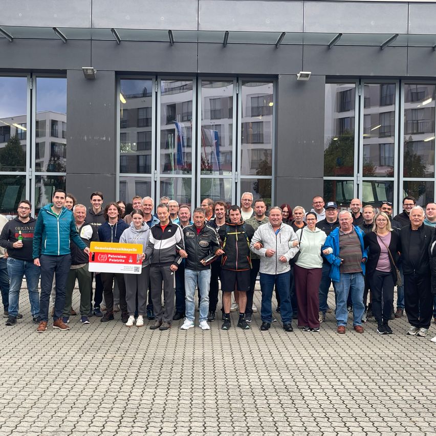Eine Gruppe von Menschen steht vor einem modernen Gebäude und posiert für ein Foto mit einem Banner. Sie tragen Freizeitkleidung und wirken fröhlich. Hinter ihnen spiegelt die Glasfassade des Gebäudes die Umgebung wider.