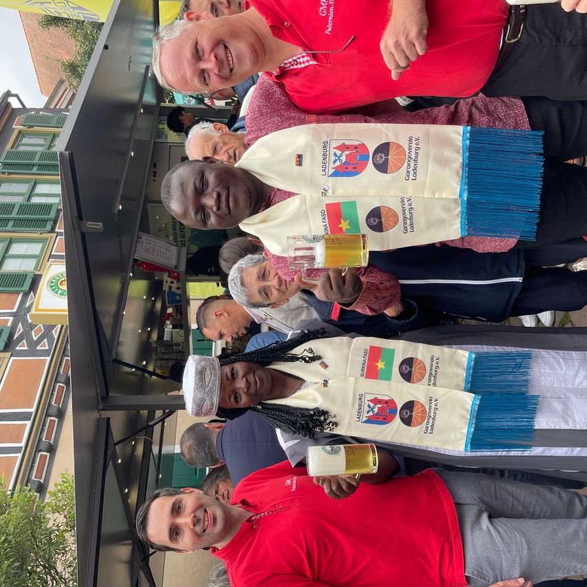 Gruppe von Menschen, die lächeln, Biergläser halten, traditionelle Schals mit Logos tragen, stehen unter einem Baldachin mit Gebäuden und Bäumen im Hintergrund.