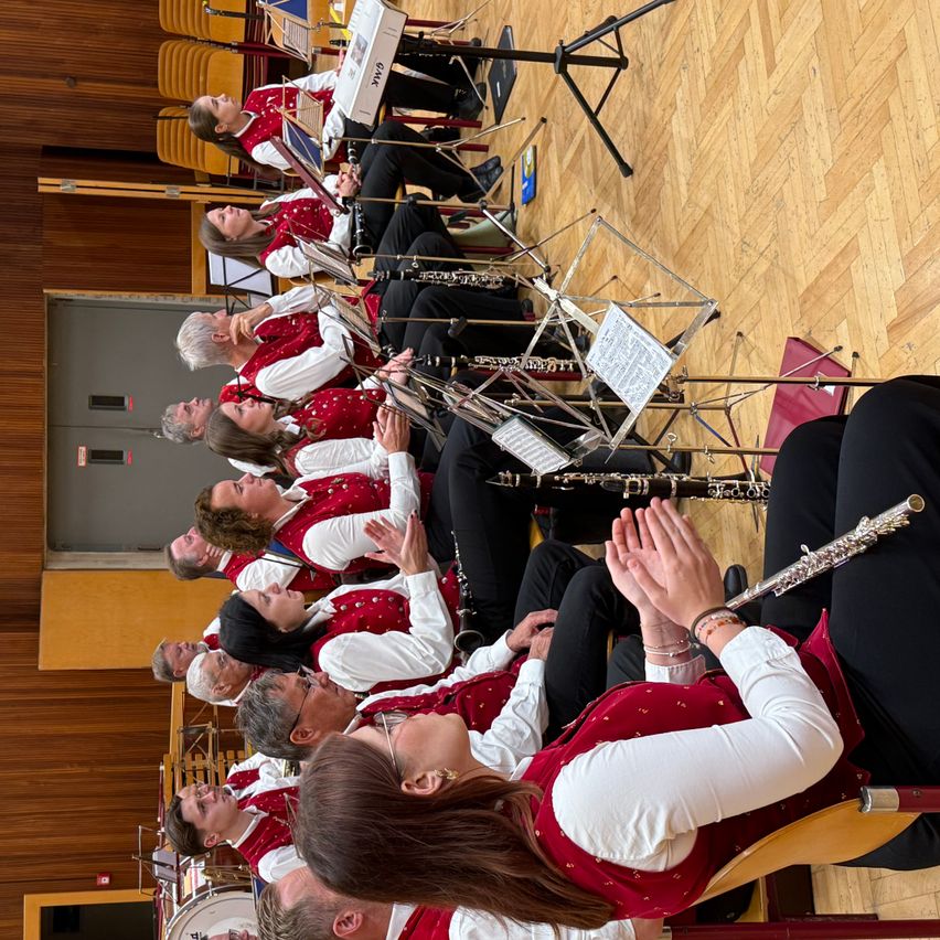 Eine Gruppe von Musikern in roten und weißen Westen spielt Klarinetten und andere Instrumente, wobei einige Mitglieder klatschen. Der Veranstaltungsort verfügt über Holzböden und eine graue Wand mit einer Tür.
