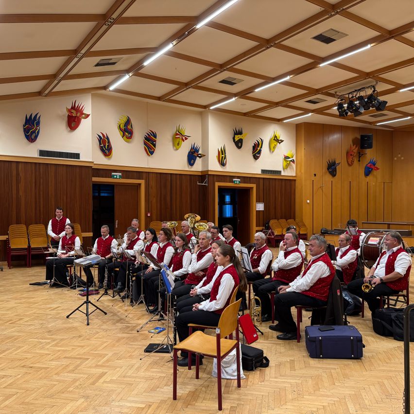 Ein Orchester sitzt in einem Raum mit Holzböden und Wänden und spielt Musikinstrumente. Mehrere Masken hängen an der Wand.