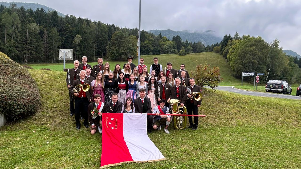Eine Gruppe von Menschen in traditioneller deutscher Kleidung posiert für ein Foto auf einem Grashügel mit Musikinstrumenten. Einige Personen halten eine Fahne, und der Hintergrund zeigt Berge.