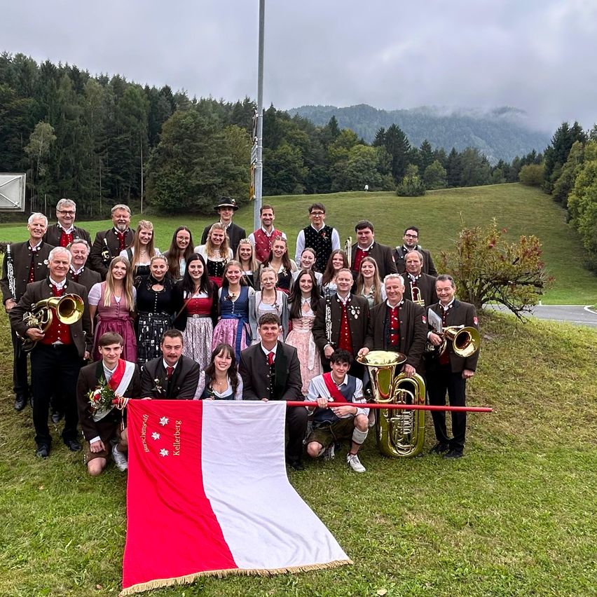Eine Gruppe von Menschen in traditioneller deutscher Kleidung posiert für ein Foto auf einem Grashügel mit Musikinstrumenten. Einige Personen halten eine Fahne, und der Hintergrund zeigt Berge.