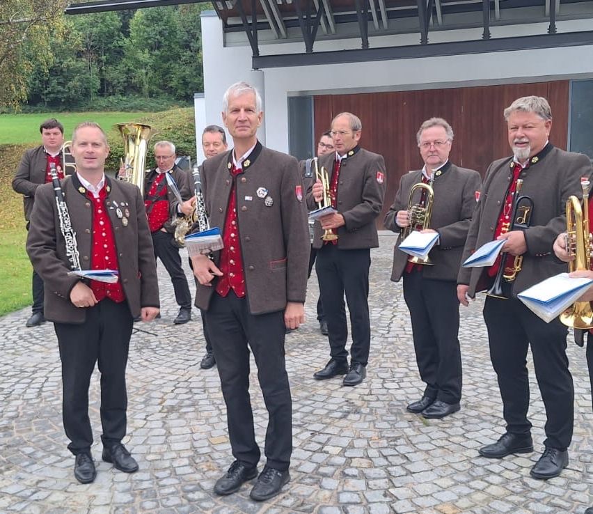 Eine Gruppe von Männern in traditionellen Uniformen, möglicherweise Musiker, stehen zusammen und halten verschiedene Musikinstrumente, darunter Trompeten, Tuben und Klarinetten. Sie lächeln und scheinen für ein Foto zu posieren.