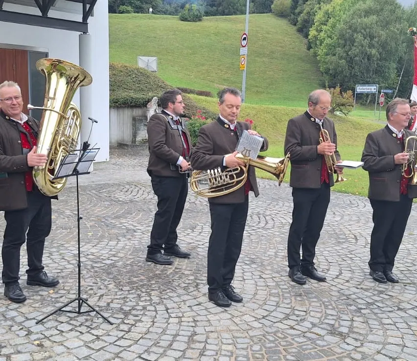 Fünf Musiker stehen auf einem Kopfsteinpflasterweg und spielen Blechblasinstrumente, einer hält ein Notenbuch. Dahinter ist ein grüner Hügel mit einem Stoppschild und einem kleinen Gebäude.
