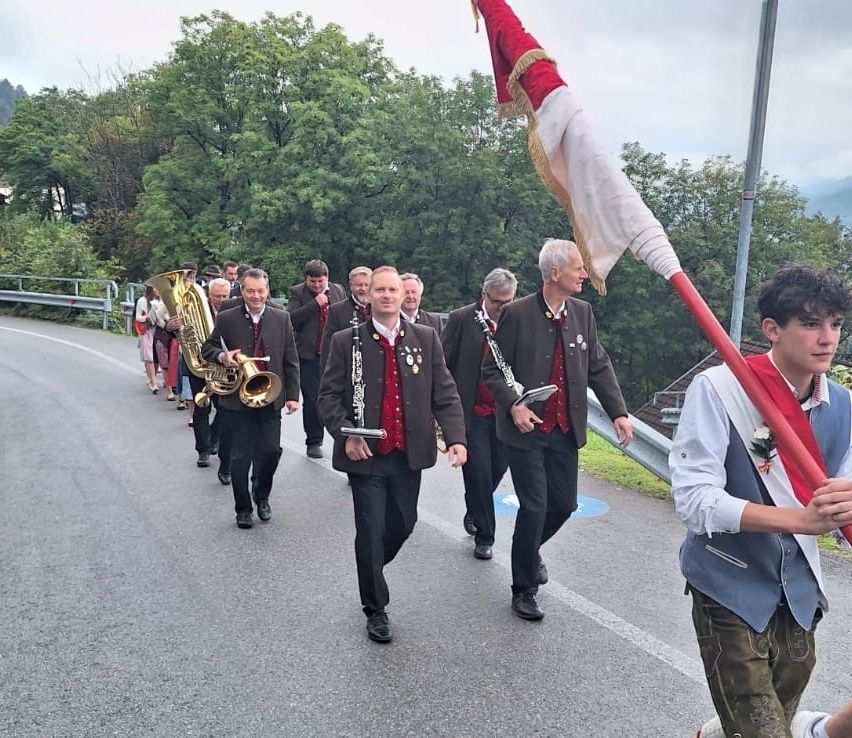 Eine Gruppe von Männern in Uniformen marschiert die Straße entlang, trägt Musikinstrumente und eine Fahne. Bäume säumen die Straße, und der Himmel ist bewölkt.