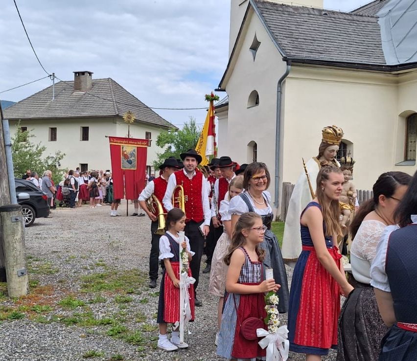 Eine Gruppe von Menschen in traditioneller bayerischer Kleidung versammelt sich vor einer Kirche. Eine Frau hält einen Blumenstrauß, während ein Mädchen eine Trompete spielt. Dahinter sind Musiker und eine Statue einer Frau zu sehen.