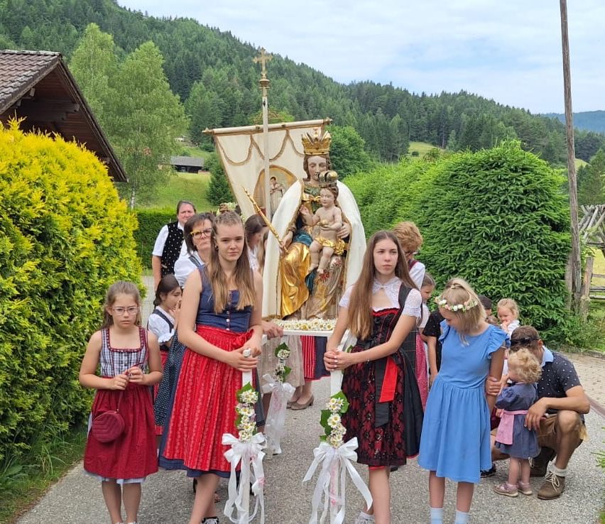 Eine Gruppe von Kindern in traditionellen Kostümen trägt eine geschmückte Kutsche mit einer Statue einer Mutter und eines Kindes. Sie sind von Grünpflanzen und einer Bergkulisse umgeben.