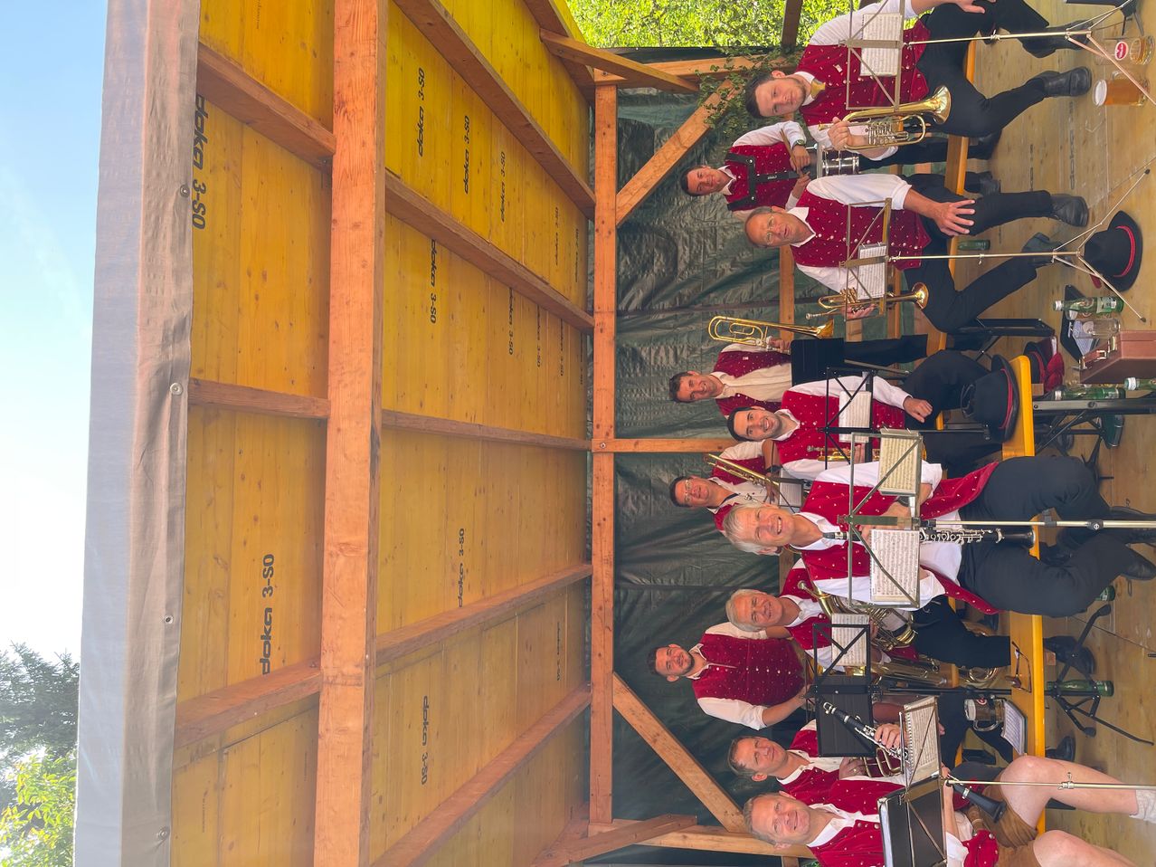Eine Gruppe von Musikern in roten und weißen Westen spielt mit Blechblasinstrumenten in einer Holzstruktur. Die Holzstruktur hat gelbe Paneele mit Text und Zahlen. Die Musiker schauen in die gleiche Richtung.