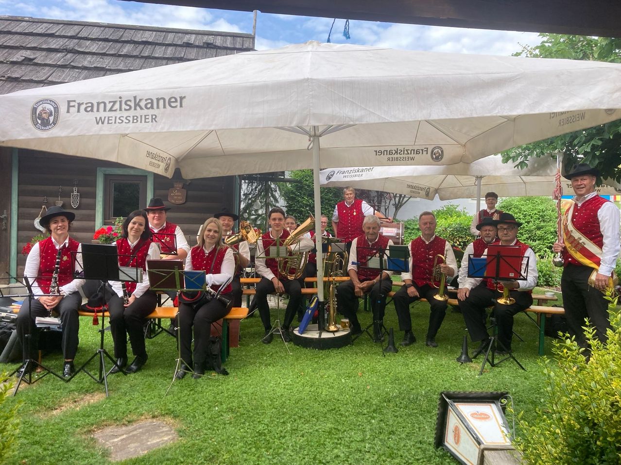 Eine Blaskapelle in traditioneller Kleidung sitzt unter weißen Schirmen auf einem Rasen, mit Musikinstrumenten und Noten. Ein Gebäude mit Tür und Fenster ist im Hintergrund.