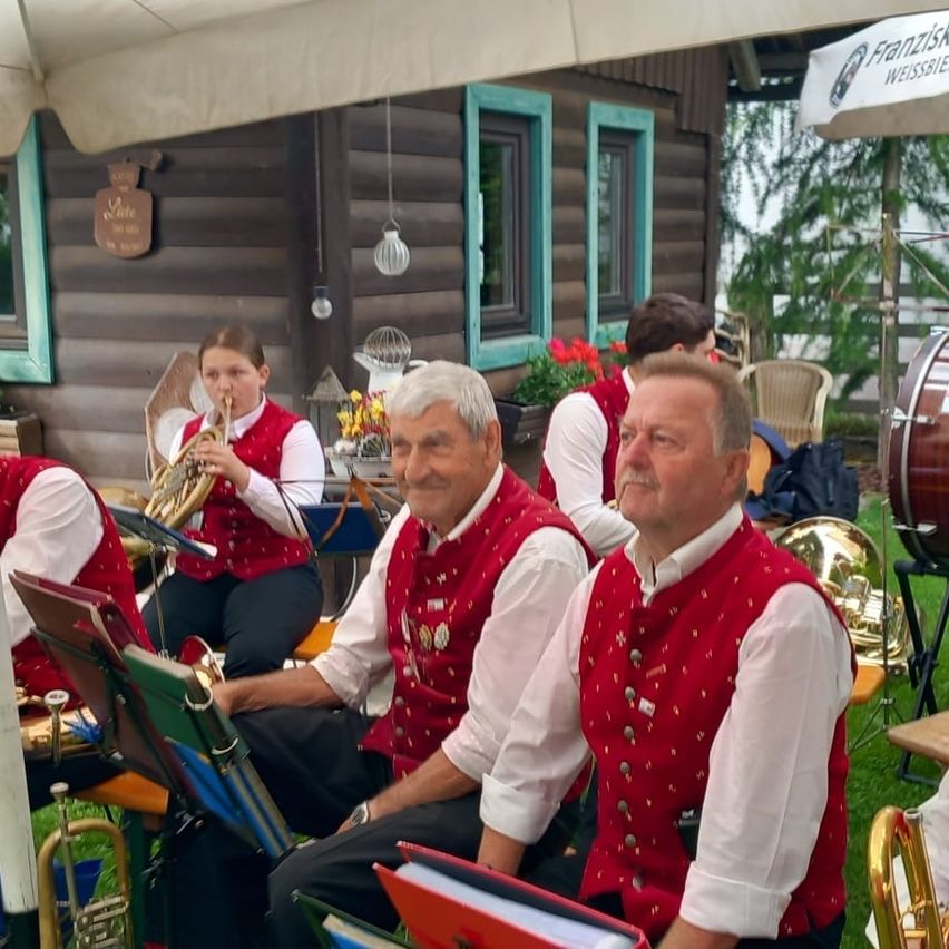 Eine Gruppe von Musikern in roten Westen sitzt auf Stühlen auf einem Rasen und spielt Instrumente. Eine Frau in einem roten Westchen spielt die Trompete. Dahinter ist ein Holzhaus mit blauer Umrandung und einem Schild zu sehen.