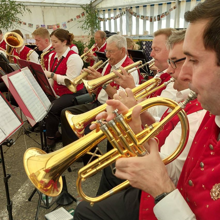 Eine Brassband spielt in einem Zelt. Sie tragen rote Westen und weiße Hemden. Sie spielen Trompeten, Posaunen und Saxophone. Sie sitzen auf Stühlen und halten Notenständer mit Notenpapieren. Das Zelt hat weiße Wände und Glasfenster mit Dekorationen.