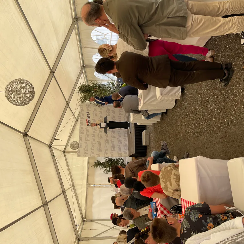 Eine Gruppe von Menschen versammelt sich in einem Zelt. Einige stehen, andere sitzen auf Stühlen um Tische herum. Zwei Personen stehen auf der Bühne, eine davon spricht. Eine Banner mit Text ist an der Wand angebracht.