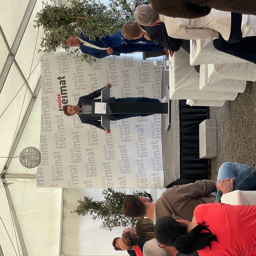 Ein Mann hält eine Rede an einem Rednerpult, während das Publikum auf Stühlen sitzt. Im Hintergrund steht der Schriftzug 'mein Heimat'.