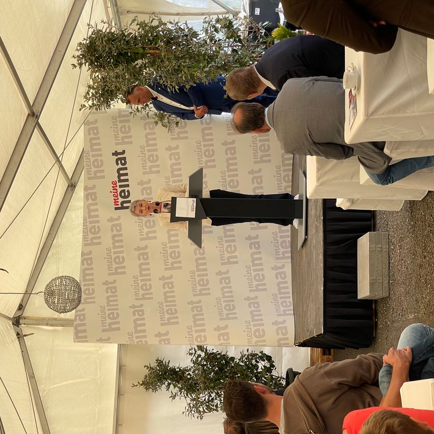 Eine Gruppe von Menschen versammelt sich unter einem Zelt, wahrscheinlich bei einer Veranstaltung. Ein Mann spricht an einem Podium, und mehrere andere hören aufmerksam zu.
