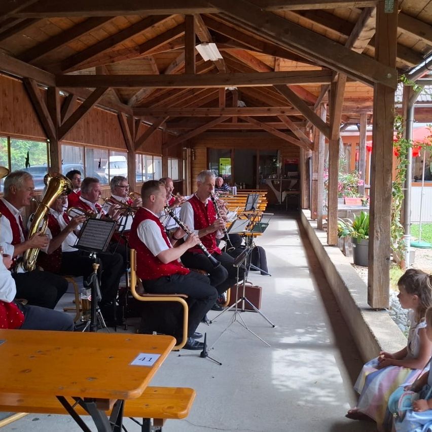 Eine Blaskapelle übt in einem Pavillon, Musiker spielen verschiedene Instrumente. Ein Mädchen beobachtet von der Seite.