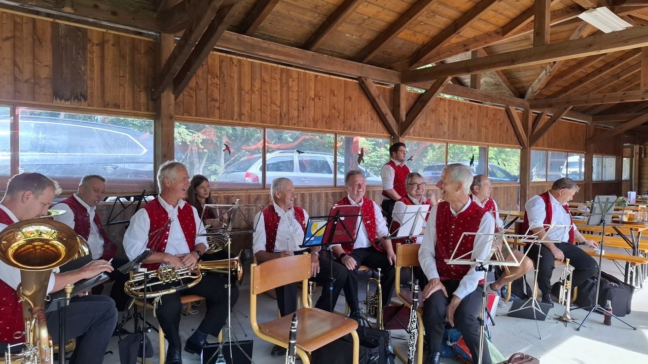 Eine Gruppe älterer Musiker in roten Westen und weißen Hemden sitzt in einem Raum mit Holzwänden und Glasfenstern. Sie spielen verschiedene Instrumente. Ein Auto ist draußen geparkt.