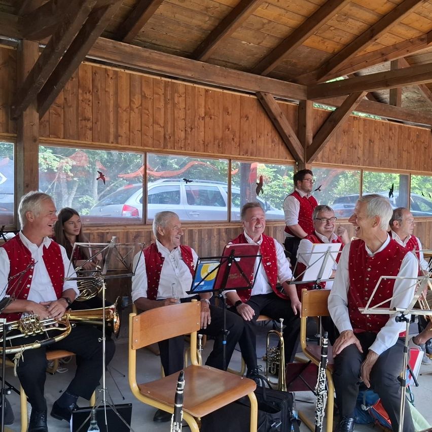 Eine Gruppe älterer Musiker in roten Westen und weißen Hemden sitzt in einem Raum mit Holzwänden und Glasfenstern. Sie spielen verschiedene Instrumente. Ein Auto ist draußen geparkt.