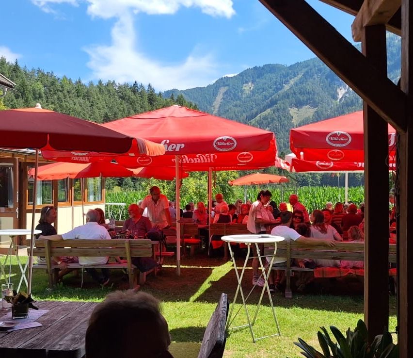Outdoor-Essbereich mit roten Sonnenschirmen, Holzbänken und Tischen. Mehrere Menschen sitzen und stehen. Berge im Hintergrund.