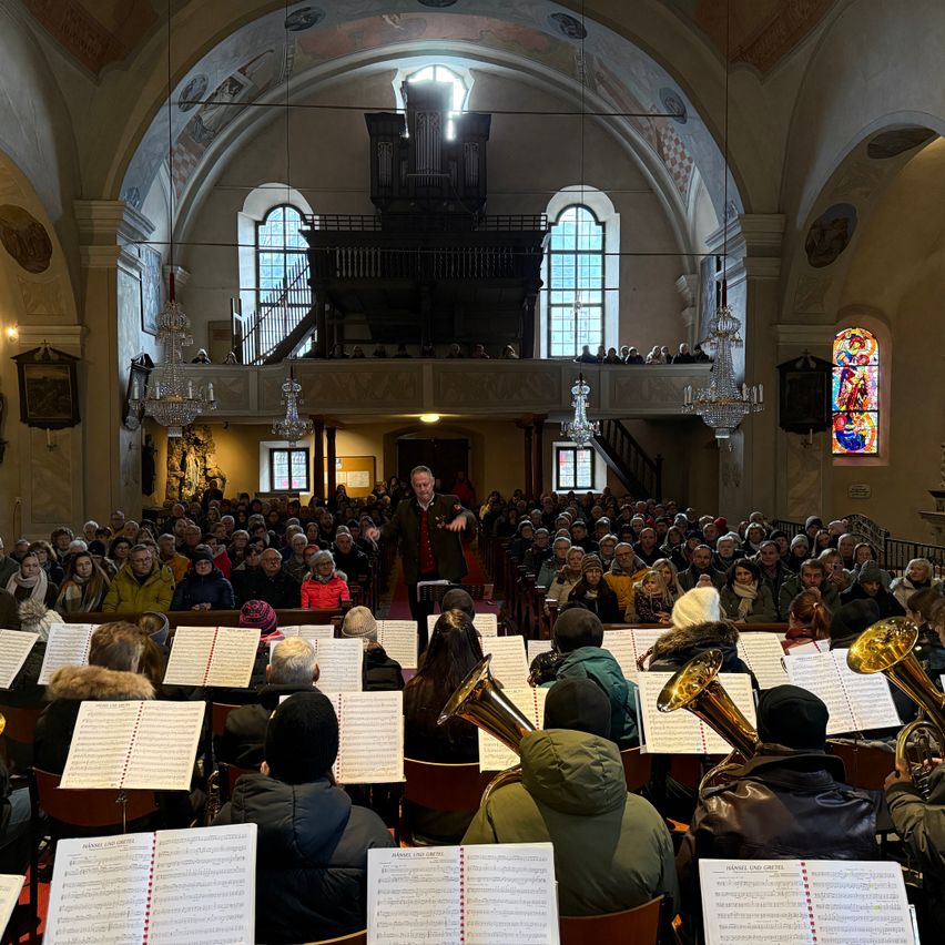 Ein großes Publikum ist in einer Kirche versammelt und beobachtet einen Dirigenten, der eine Aufführung mit einem Blechbläserensemble leitet. Das Publikum sitzt auf Bänken, und die Musiker stehen vor Notenständern.