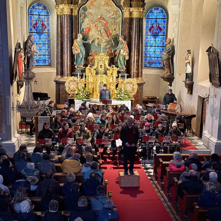 Ein Musikensemble spielt in einer Kirche, wobei der Dirigent auf einer Plattform vor dem Orchester steht. Die Gemeinde sitzt auf beiden Seiten des Mittelgangs.