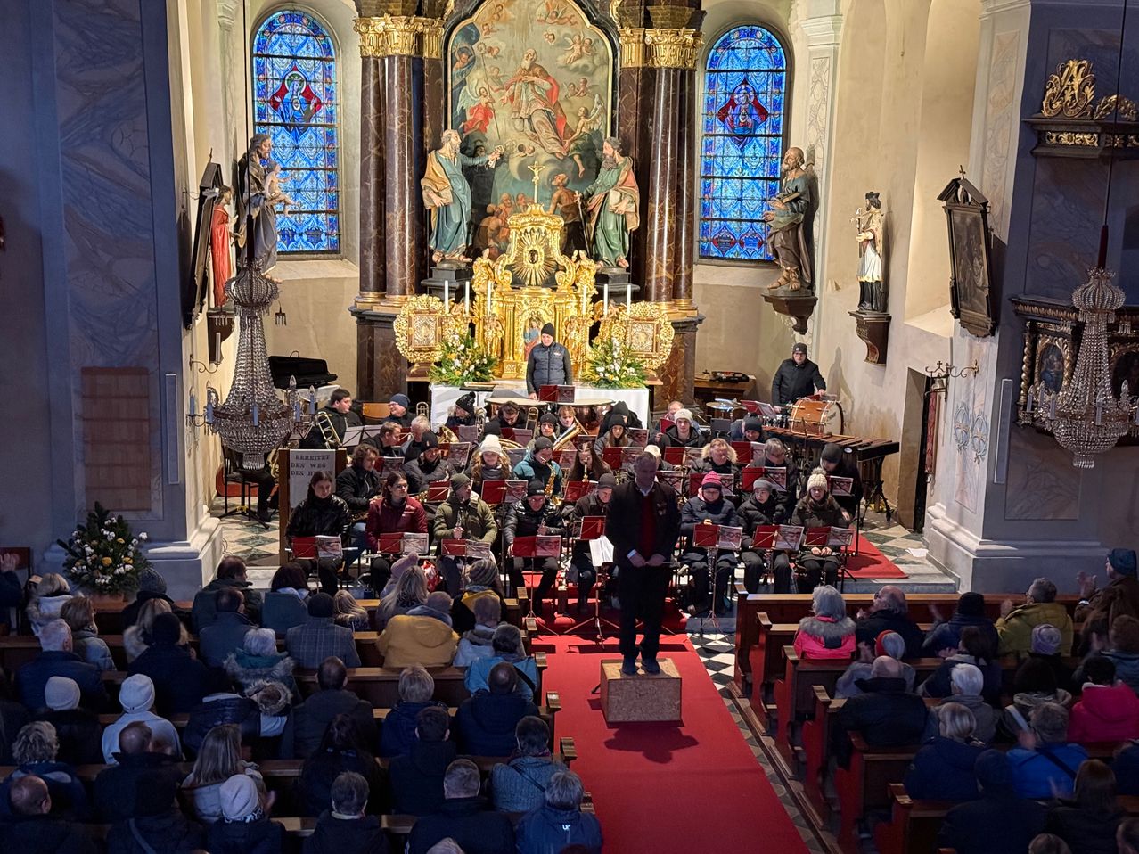 Ein Musikensemble spielt in einer Kirche, wobei der Dirigent auf einer Plattform vor dem Orchester steht. Die Gemeinde sitzt auf beiden Seiten des Mittelgangs.