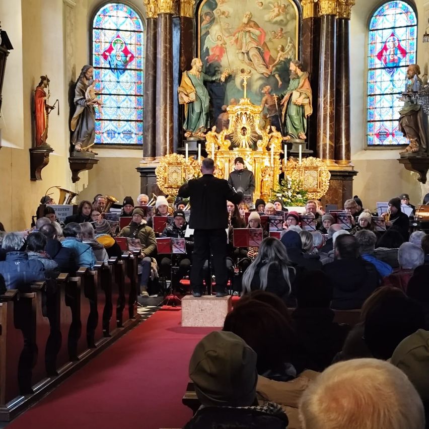Ein Dirigent leitet eine Band in einer Kirche mit Buntglasfenstern, einem großen Gemälde und Statuen. Das Publikum sitzt auf Kirchenbänken und beobachtet die Aufführung.