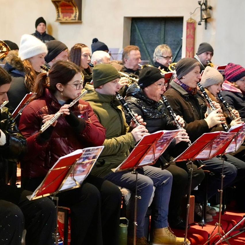 Eine Gruppe von Musikern sitzt in einer Reihe und spielt ihre Instrumente, darunter Flöten, Klarinetten und Trompeten. Sie tragen Winterkleidung und einige von ihnen haben Brillen. Sie spielen wahrscheinlich bei einem Konzert.