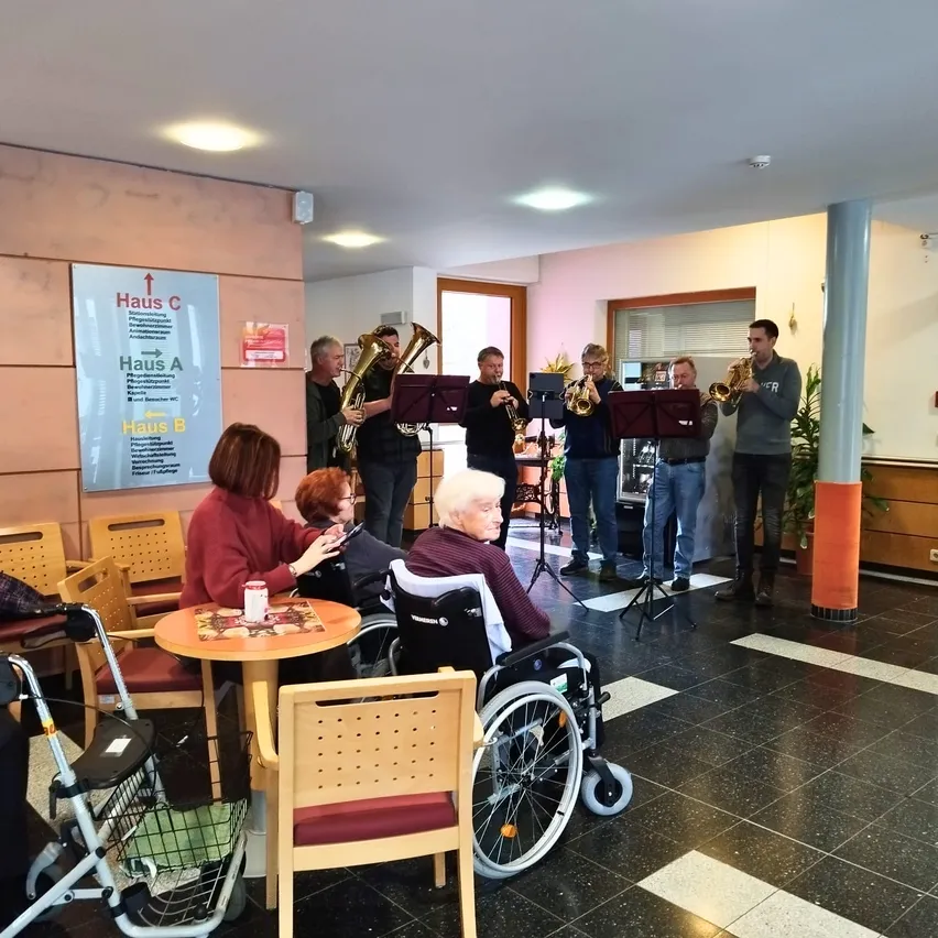 Eine Gruppe von Musikern in einem Flur spielt Instrumente, während einige Menschen in Rollstühlen im Vordergrund sitzen. Ein Schild an der Wand zeigt die Richtung zu verschiedenen Häusern.