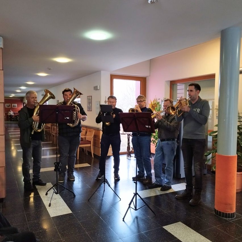 Sechs Männer spielen Blechblasinstrumente in einem Raum. Sie stehen in der Nähe von Notenständern mit Musikbüchern. Der Raum hat einen gefliesten Boden und weiße Wände. Es gibt Topfpflanzen und Holzbänke im Raum.