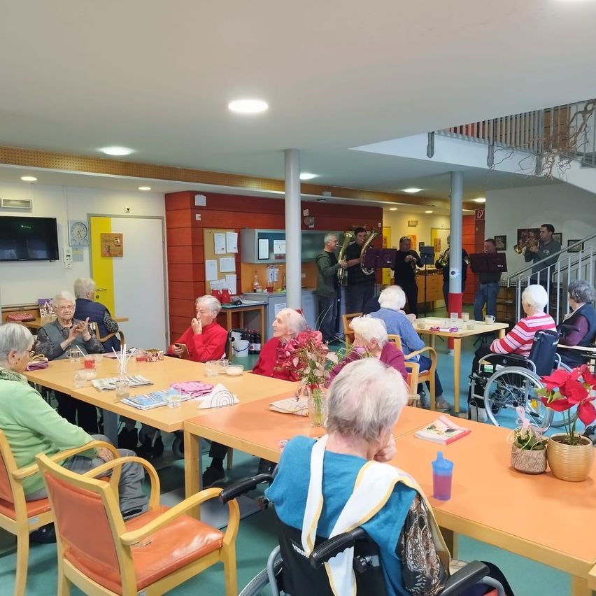 Eine Gruppe älterer Menschen sitzt um Tische herum und genießt eine musikalische Aufführung einer Band. Der Raum ist hell und die Musiker befinden sich auf einer höheren Ebene. Blumen und Tassen sind auf den Tischen.