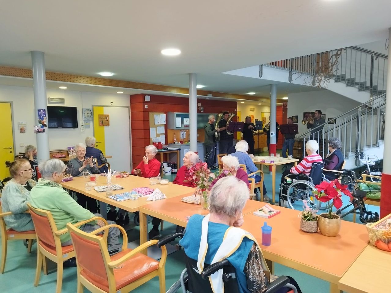 Eine Gruppe älterer Menschen sitzt um Tische herum und genießt eine musikalische Aufführung einer Band. Der Raum ist hell und die Musiker befinden sich auf einer höheren Ebene. Blumen und Tassen sind auf den Tischen.