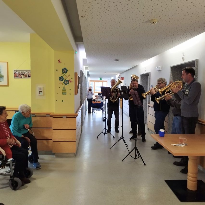 Eine Gruppe von Musikern spielt Trompeten in einem Flur eines Pflegeheims. Mehrere ältere Menschen sitzen in Rollstühlen und beobachten die Aufführung.