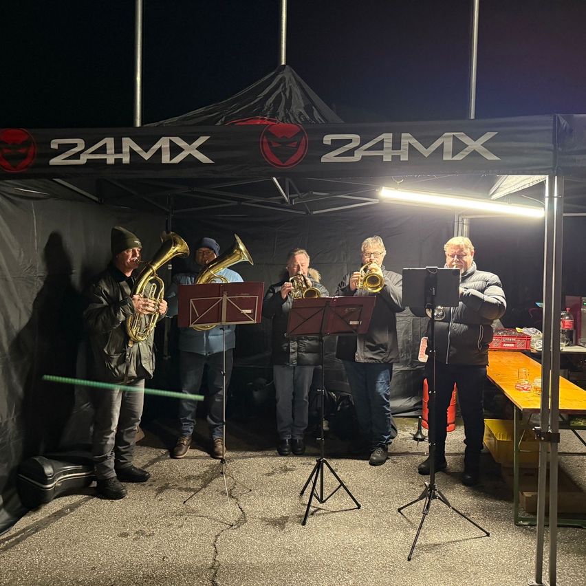 Fünf Männer spielen Trompeten unter einem Zelt mit dem Logo 24MX. Sie sind von einer Lampe beleuchtet. Ein Tisch mit Getränken befindet sich rechts.