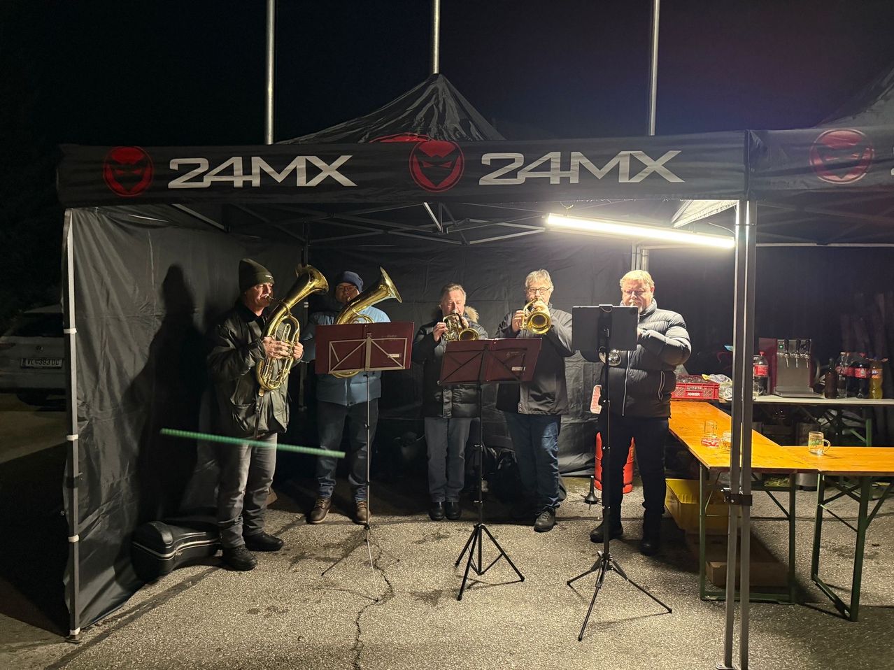 Fünf Männer spielen Trompeten unter einem Zelt mit dem Logo 24MX. Sie sind von einer Lampe beleuchtet. Ein Tisch mit Getränken befindet sich rechts.