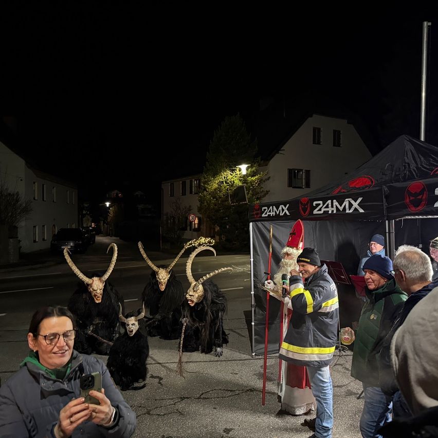 Eine Menschenmenge versammelt sich bei Nacht. Drei Personen mit Masken und Hörnern sitzen auf der Straße. Ein Mann in einem Weihnachtsmannkostüm hält ein Mikrofon und spricht.